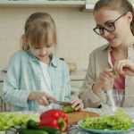 Adorable little girl is helping mother in kitchen cutting vegetables for salad and talking making dinner together. Healthy nutrition, lifestyle and people concept.